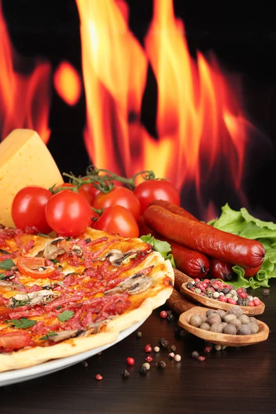 Stock image Delicious pizza, salami, tomatoes and spices on wooden table on flame background