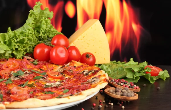 Deliciosa pizza, salami, tomates y especias en mesa de madera sobre fondo de llama — Foto de Stock