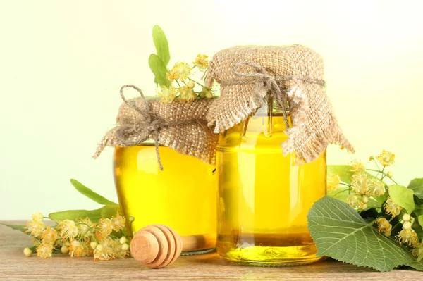 Jars with linden honey and flowers on wooden table on green background — Stock Photo, Image
