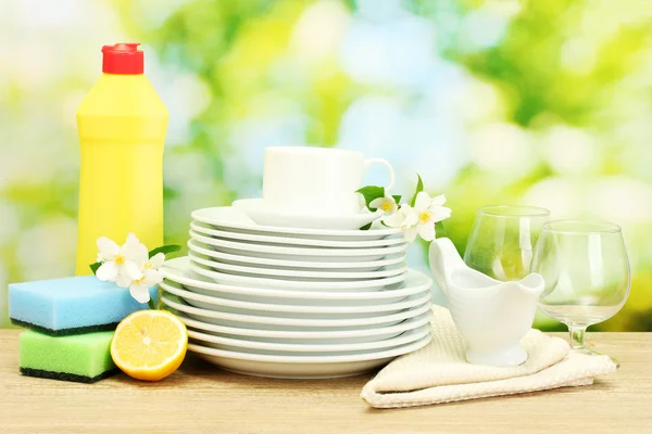 Platos limpios vacíos, vasos y tazas con líquido lavavajillas, esponjas y limón sobre mesa de madera sobre fondo verde —  Fotos de Stock