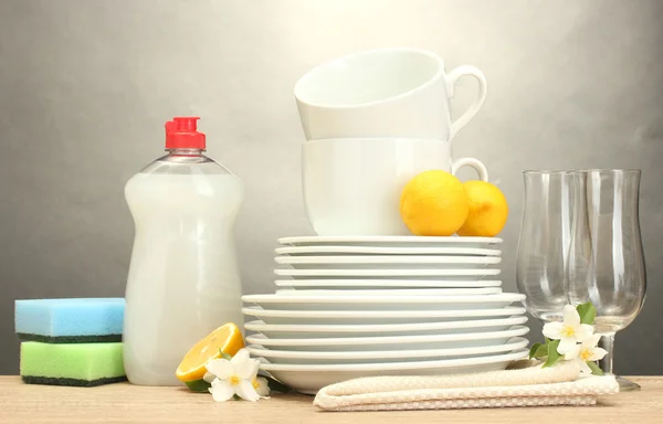 Piatti puliti vuoti, bicchieri e tazze con detersivo per piatti, spugne e limone su tavolo in legno su fondo grigio — Foto Stock