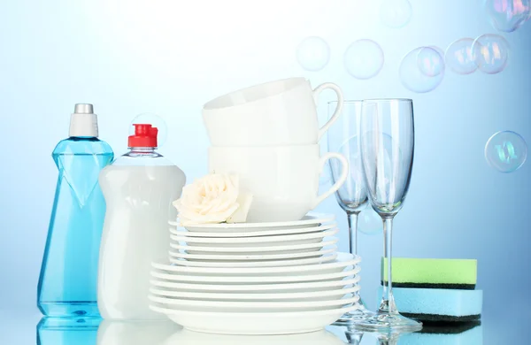 Platos limpios vacíos, vasos y tazas con líquido lavavajillas y esponjas sobre fondo azul — Foto de Stock