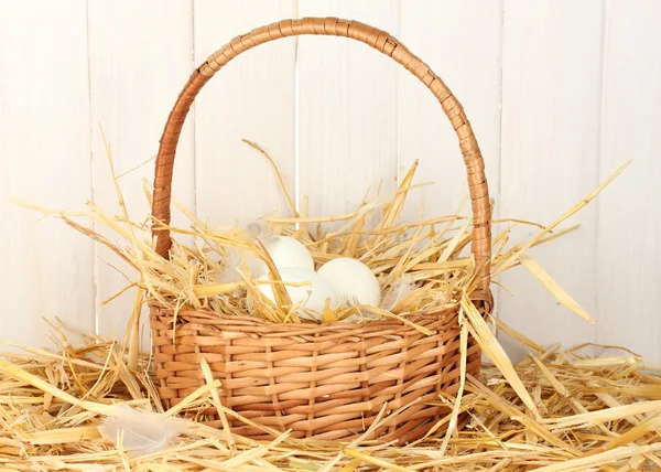 Ovos brancos em uma cesta de vime na palha no fundo de madeira branco — Fotografia de Stock