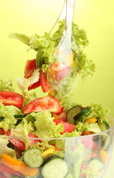 Салат из свежих овощей в прозрачной миске с ложкой и вилкой на зеленом фоне — стоковое фото