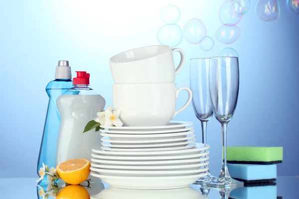 Pratos limpos vazios, copos e copos com líquido de lavar louça, esponjas e limão sobre fundo azul — Fotografia de Stock