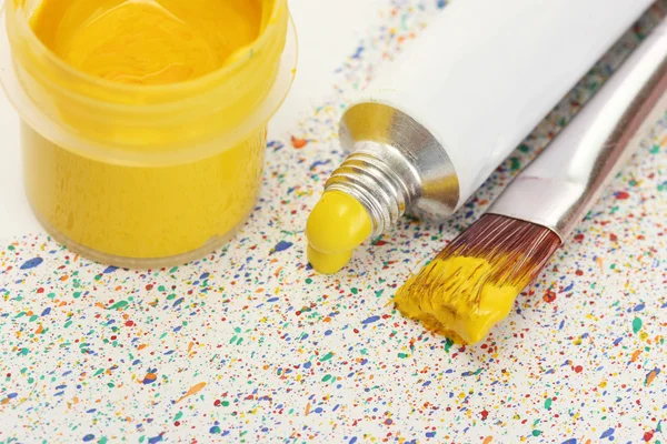 Brushes with colorful paint, tube with watercolor and jar with gouache on colorful splashes background close-up — Stock Photo, Image
