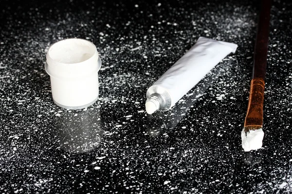 Glas mit weißer Gouache, Pinsel und Tube mit weißem Aquarell auf schwarzem Hintergrund, mit weißer Farbe bespritzt Nahaufnahme — Stockfoto