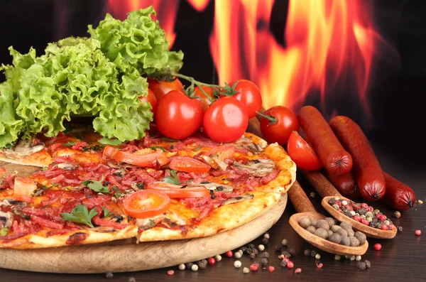 Heerlijke pizza, salami, tomaten en kruiden op houten tafel op vlam achtergrond — Stockfoto