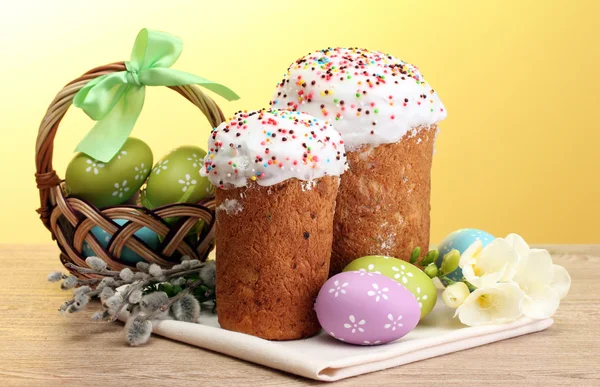 Hermosos pasteles de Pascua, huevos coloridos en cesta y ramitas de sauce en la mesa de madera sobre fondo amarillo —  Fotos de Stock