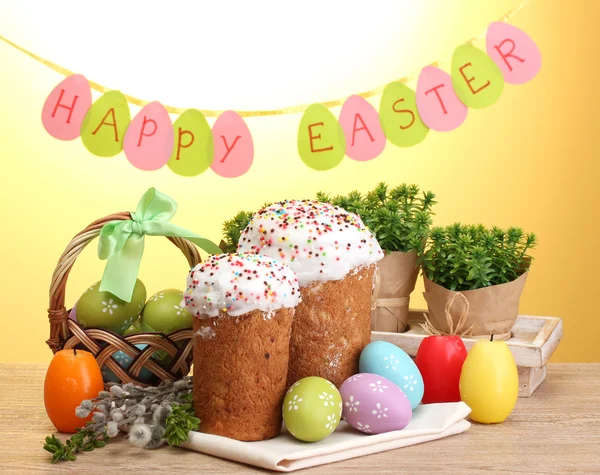 Kaunis pääsiäiskakut, värikkäät munat korissa ja kynttilät puupöydällä keltaisella taustalla — kuvapankkivalokuva