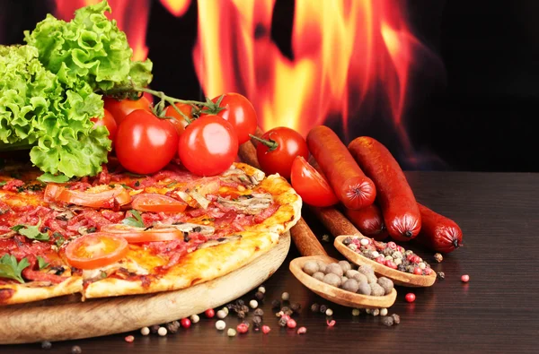 stock image Delicious pizza, salami, tomatoes and spices on wooden table on flame background