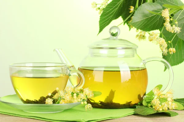 Чайник и чашка с липовым чаем и цветами на деревянном столе на зеленом фоне — стоковое фото