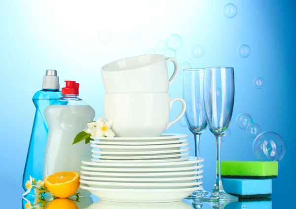 Puste czyste talerze, szklanki i kubki z płynu do mycia naczyń, gąbki i cytryny na niebieskim tle — Zdjęcie stockowe