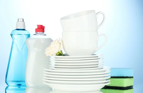 Platos y tazas limpias vacías con líquido lavavajillas y esponjas sobre fondo azul —  Fotos de Stock
