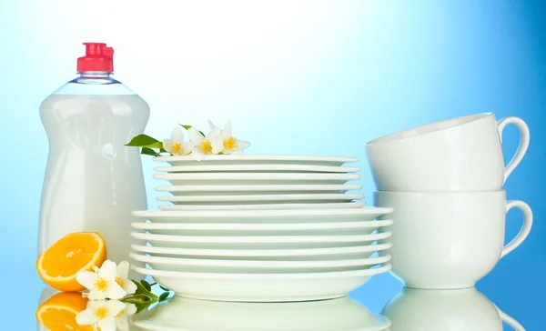 Üres tiszta tányérok, poharak és csészék és folyékony mosogatószert és a citrom, a kék háttér — Stock Fotó