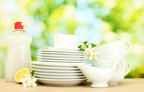 Empty clean plates and cups with dishwashing liquid, flowers and lemon on wooden table on green background — Stock Photo, Image