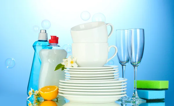 Piatti puliti vuoti, bicchieri e tazze con liquido di lavaggio piatti, spugne e limone su sfondo blu — Foto Stock