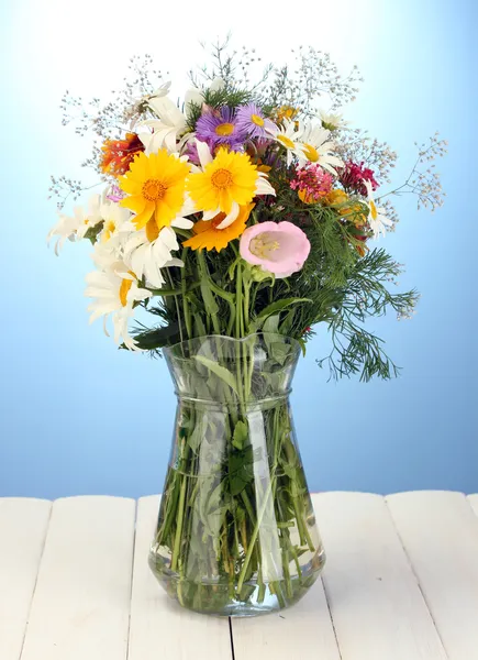 Beautiful bouquet of bright wildflowers in glass vase, on wooden table on blue background — Stock Photo, Image