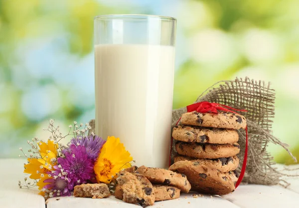 Glas Milch, Schokoladenkekse mit roter Schleife und Wildblumen auf Holztisch auf grünem Hintergrund — Stockfoto