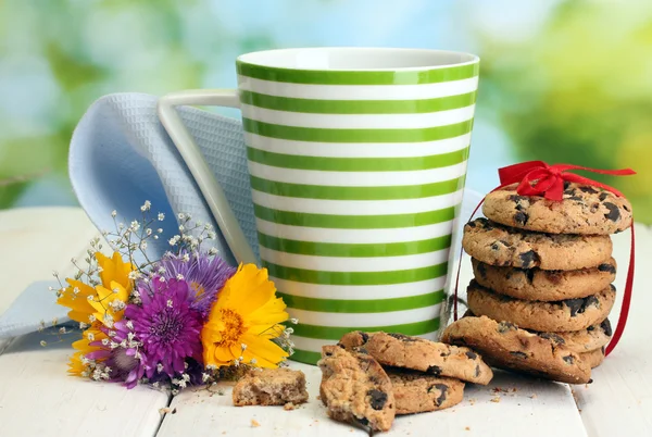 Cup of milk, chocolate chips cookies with red ribbon and wildflowers on wooden table on green background — Stock Photo, Image