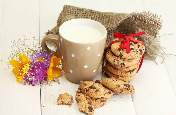 Šálek mléka, čokoládová čipy cookies s červenou stužku a květy na dřevěný stůl na zeleném pozadí — Stock fotografie
