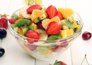 Glass bowl with fresh fruits salad and berries on white wooden table clipart