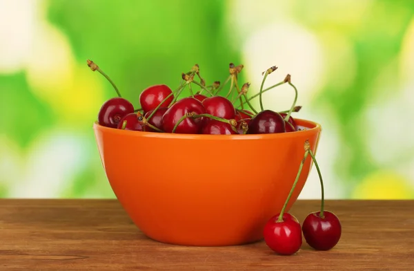Cherry in orange bowl on wooden table on green background — Stock Photo, Image