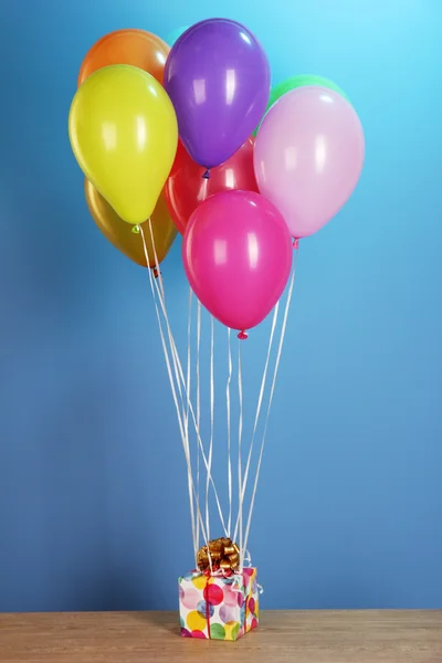 Balões coloridos segurando um presente em uma mesa de madeira em um fundo azul — Fotografia de Stock