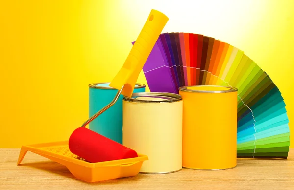Latas con pintura, rodillo, pinceles y paleta de colores sobre tabla de madera sobre fondo amarillo —  Fotos de Stock