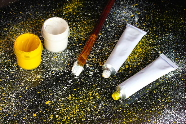 Jars with white and yellow gouache, brush and tubes with white and yellow watercolor on black background, spattered with white and yellow paint close-up — Stock Photo, Image