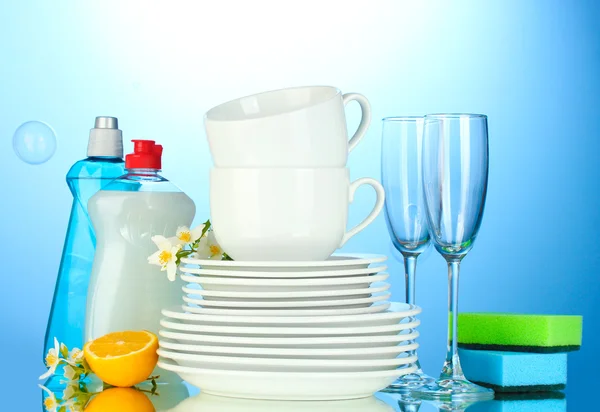 Platos limpios vacíos, vasos y tazas con líquido lavavajillas, esponjas y limón sobre fondo azul —  Fotos de Stock