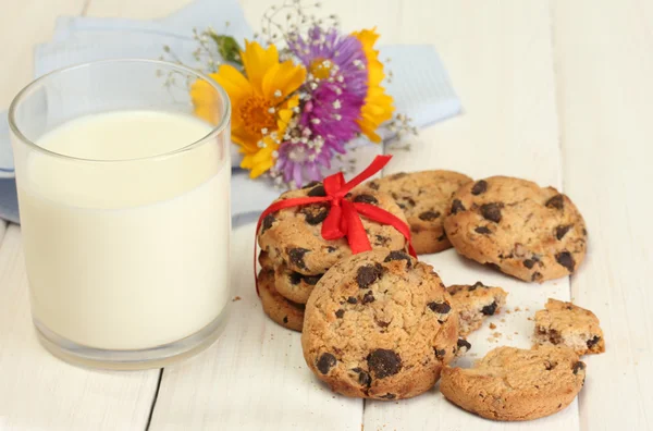 Glass of milk, chocolate chips cookies with red ribbon and wildflowers on wooden table — Stock Photo, Image