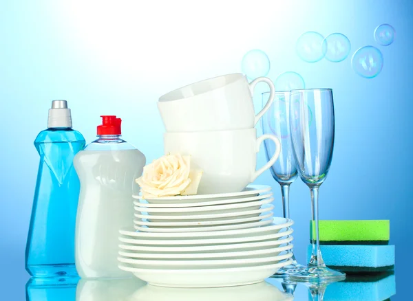 Platos limpios vacíos, vasos y tazas con líquido lavavajillas y esponjas sobre fondo azul — Foto de Stock