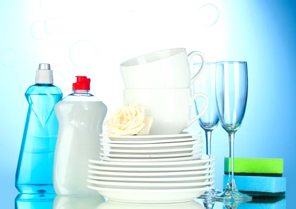 Пустые чистые тарелки, стаканы и чашки с жидкостью для мытья посуды и губки на синем фоне — стоковое фото