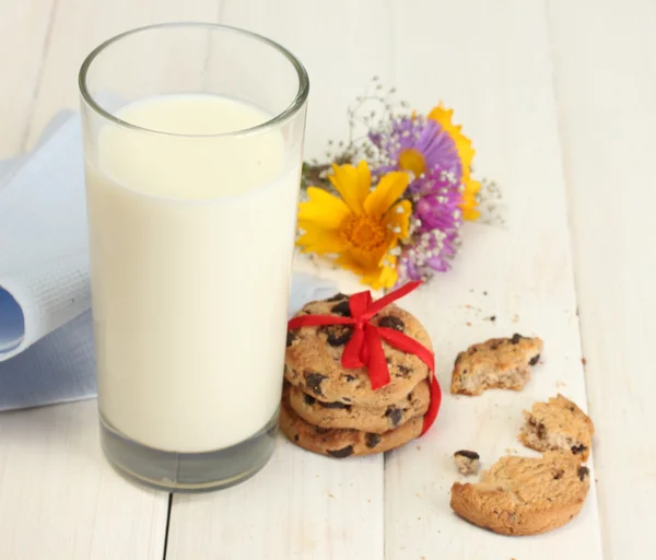 Glass of milk, chocolate chips cookies with red ribbon and wildflowers on wooden table — Stock Photo, Image