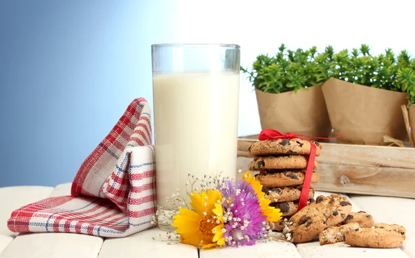 Vetro di latte, gocce di cioccolato biscotti con nastro rosso e fiori di campo su tavolo di legno su sfondo blu — Foto Stock