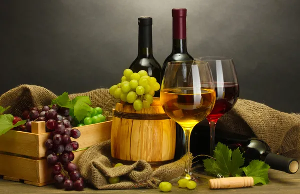 राखाडी पार्श्वभूमीवर लाकडी टेबलवर बॅरल, बाटल्या आणि वाईनचे चष्मा आणि परिपक्व द्राक्ष स्टॉक फोटो