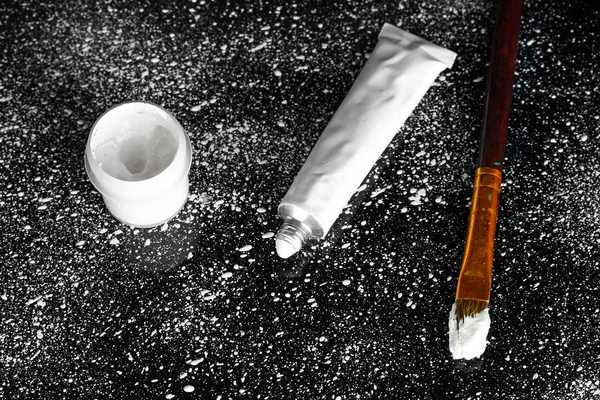 Банка с белым гуашь, кисть и трубка с белым акварелью на черном фоне, брызги белой краской крупным планом — стоковое фото