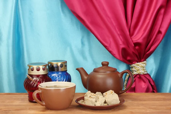 Чайник с чашкой и блюдцем со сладкой халвой на деревянном столе на фоне занавеса крупным планом — стоковое фото