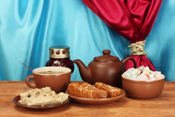 Teekanne mit Tasse und Untertassen mit orientalischen Süßigkeiten - Serbet, Halva und türkisches Vergnügen auf einem Holztisch vor dem Hintergrund eines Vorhangs in Nahaufnahme — Stockfoto
