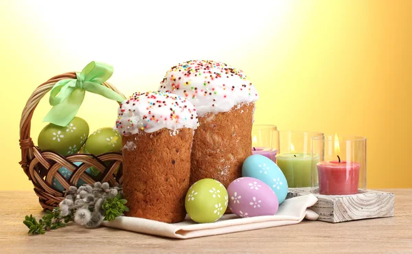 Schöne Osterkuchen, bunte Eier im Korb und Kerzen auf Holztisch auf gelbem Hintergrund — Stockfoto