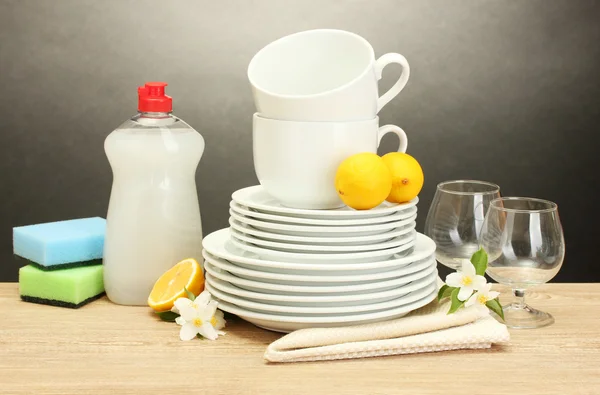 Platos limpios vacíos, vasos y tazas con líquido lavavajillas, esponjas y limón sobre mesa de madera sobre fondo gris —  Fotos de Stock