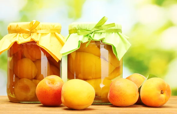 Canned apricots in a jars and sweet apricots on wooden table on green background — Stock Photo, Image
