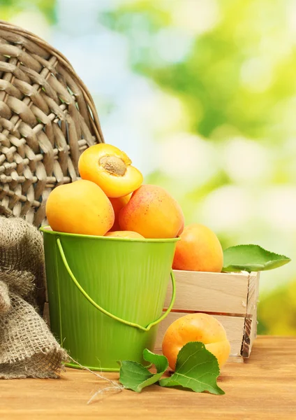 Bucket and wooden box with ripe apricots on wooden table on green background — Stock Photo, Image