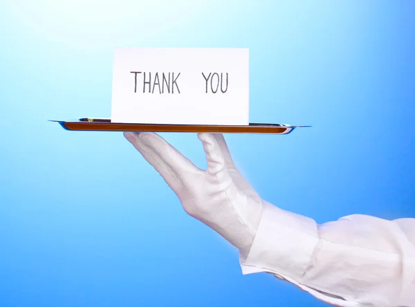Guante de mano sosteniendo bandeja de plata con tarjeta diciendo gracias sobre fondo azul — Foto de Stock