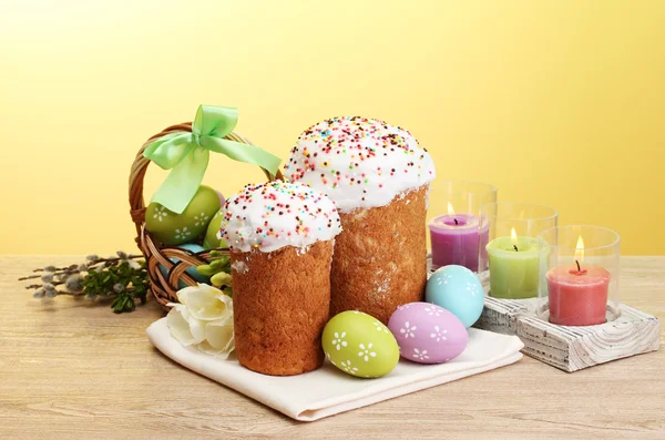 Szép húsvéti sütemény, színes tojást a kosárba, és a fából készült asztal a sárga háttér gyertyák — Stock Fotó