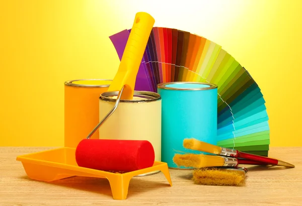Latas de estanho com tinta, rolo, escovas e paleta brilhante de cores na mesa de madeira no fundo amarelo — Fotografia de Stock