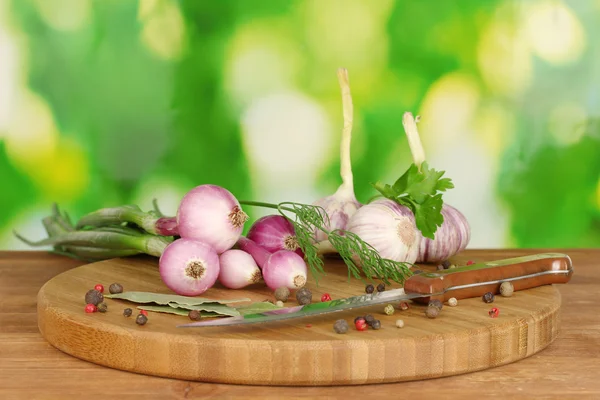 Młody czosnek i cebula na deska do krojenia z zielenią, pieprz i nóż na drewnianym stole na zielono — Zdjęcie stockowe