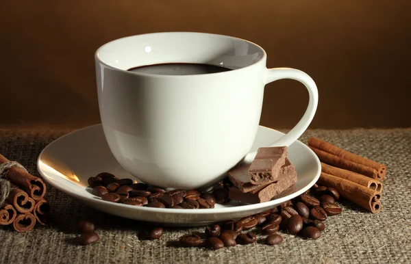 Kopp kaffe och bönor, kanelstänger och choklad på säckväv på brun bakgrund — Stockfoto