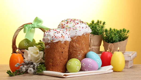 Beautiful Easter cakes, colorful eggs in basket and candles on wooden table on yellow background — Stock Photo, Image